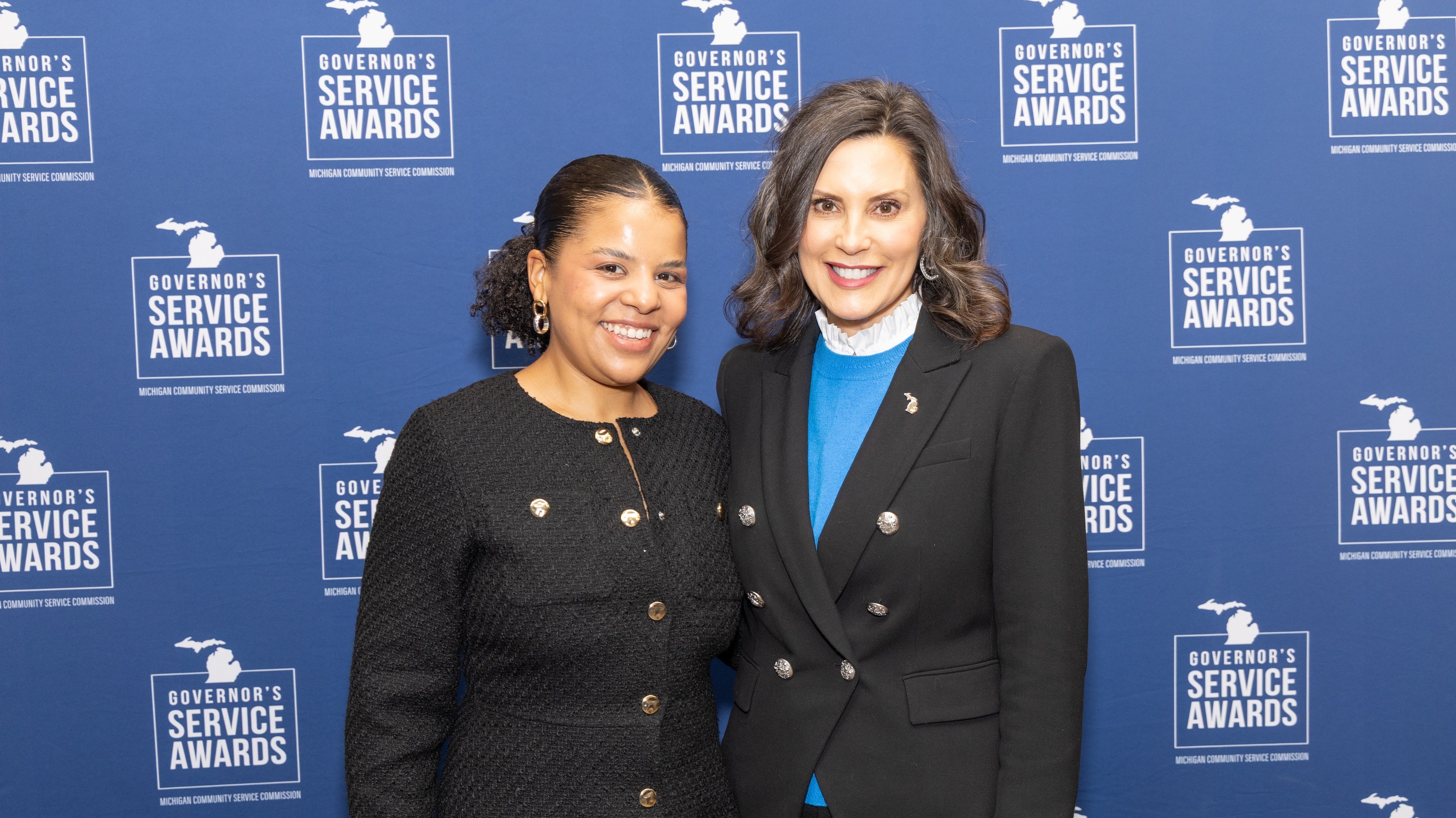  Two people standing in front of a step-and-repeat blue wall with a logo that says Governor&rsquo;s Service Awards.&nbsp;