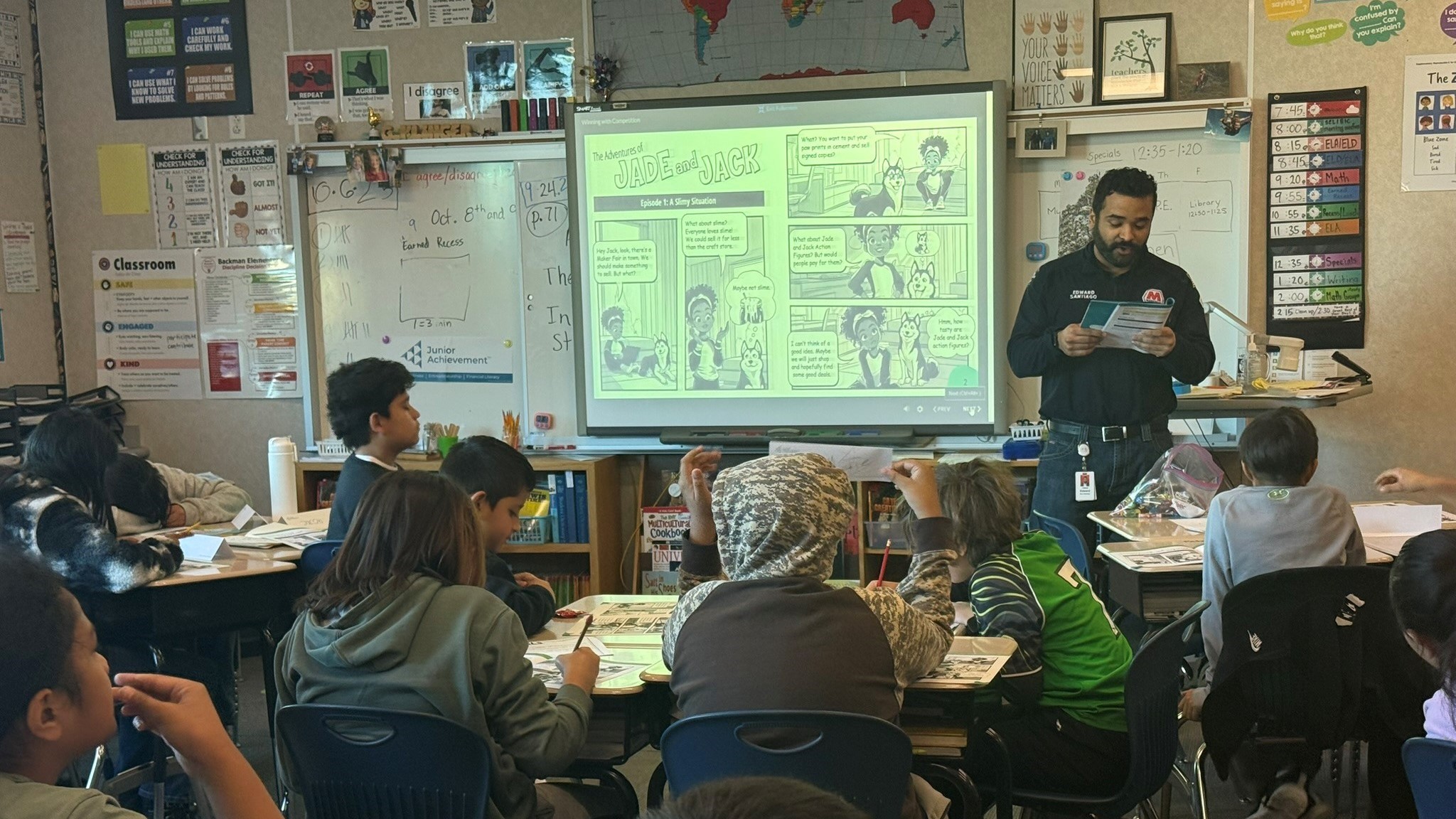 A man stands and reads to children who are seated at desks.