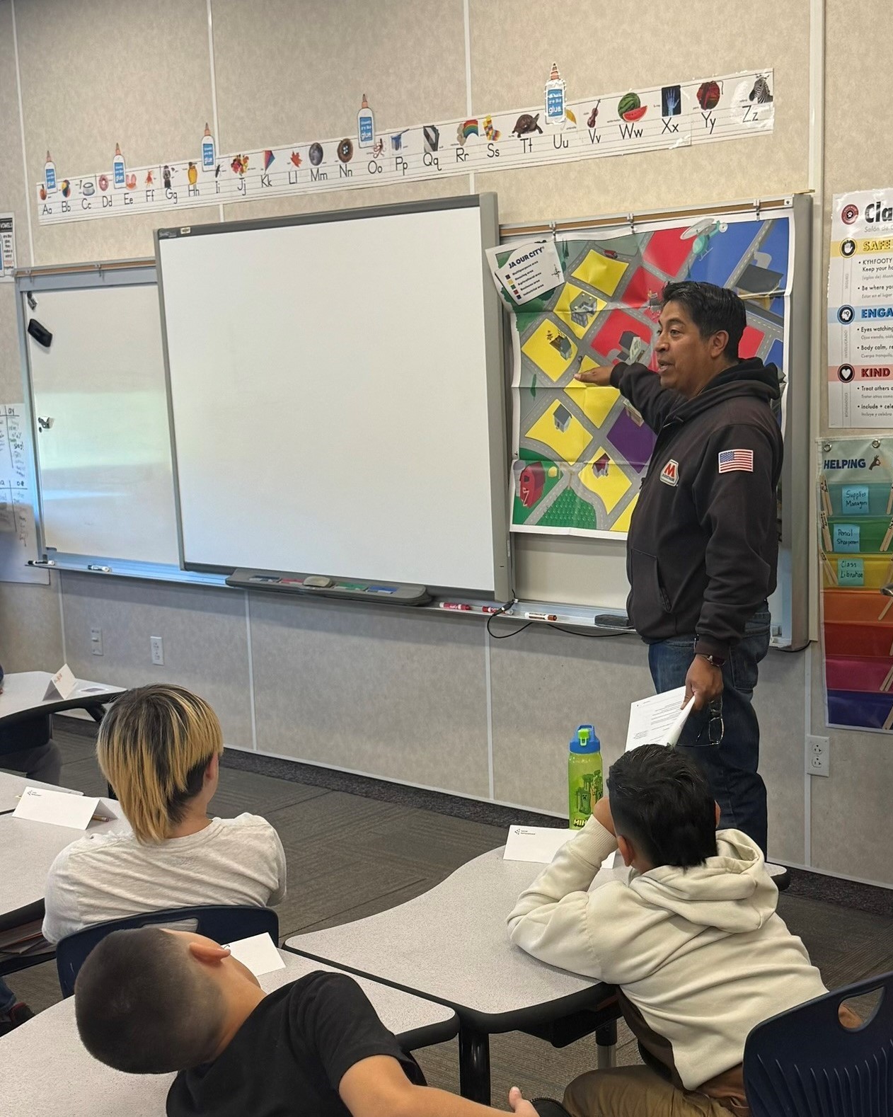 A man stands and points to a map in a classroom with children seated at desks.&nbsp;