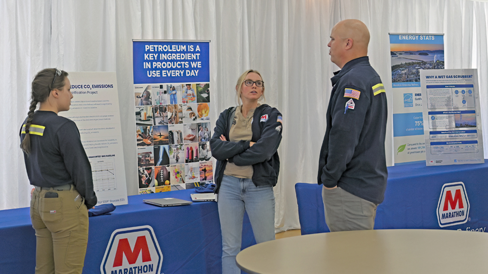 Three people talk near a table with informational posters displayed.