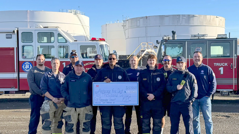 A group of people holding a large symbolic check for $10,000 in front of two fire trucks and terminal storage tanks. 