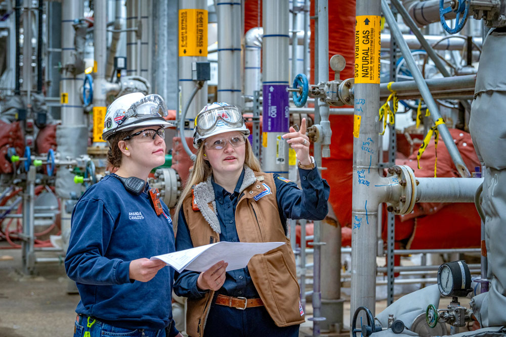 Two people with hard hats and safety gear hold a piece of paper in a unit at the refinery. 