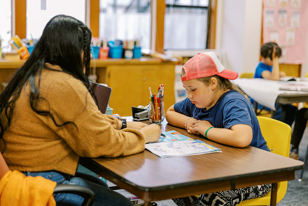 : A student sits at a table across from an adult pointing at school work.