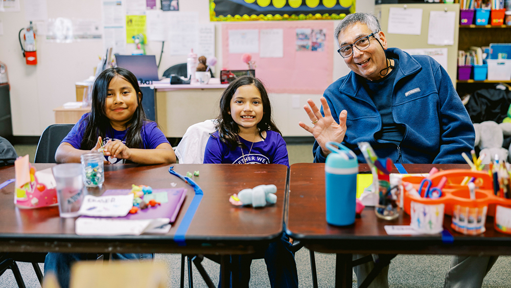 A man waves and sits next to two smiling children at an arts and crafts table.