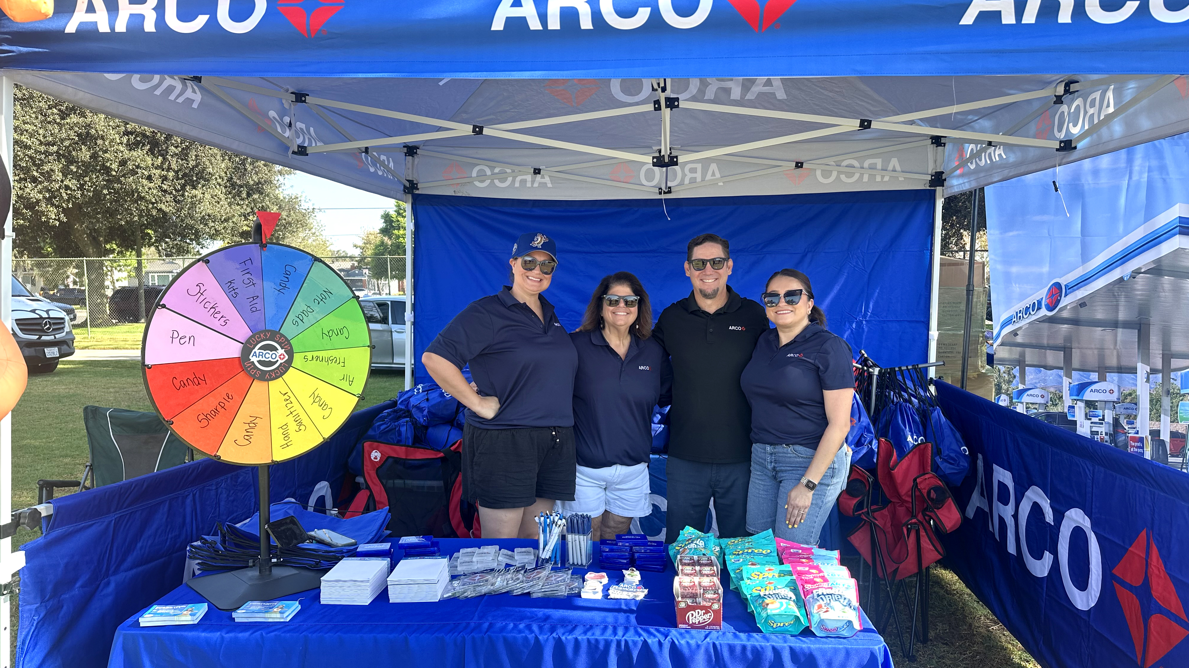 Four people standing under an ARCO branded tent with items on a table.