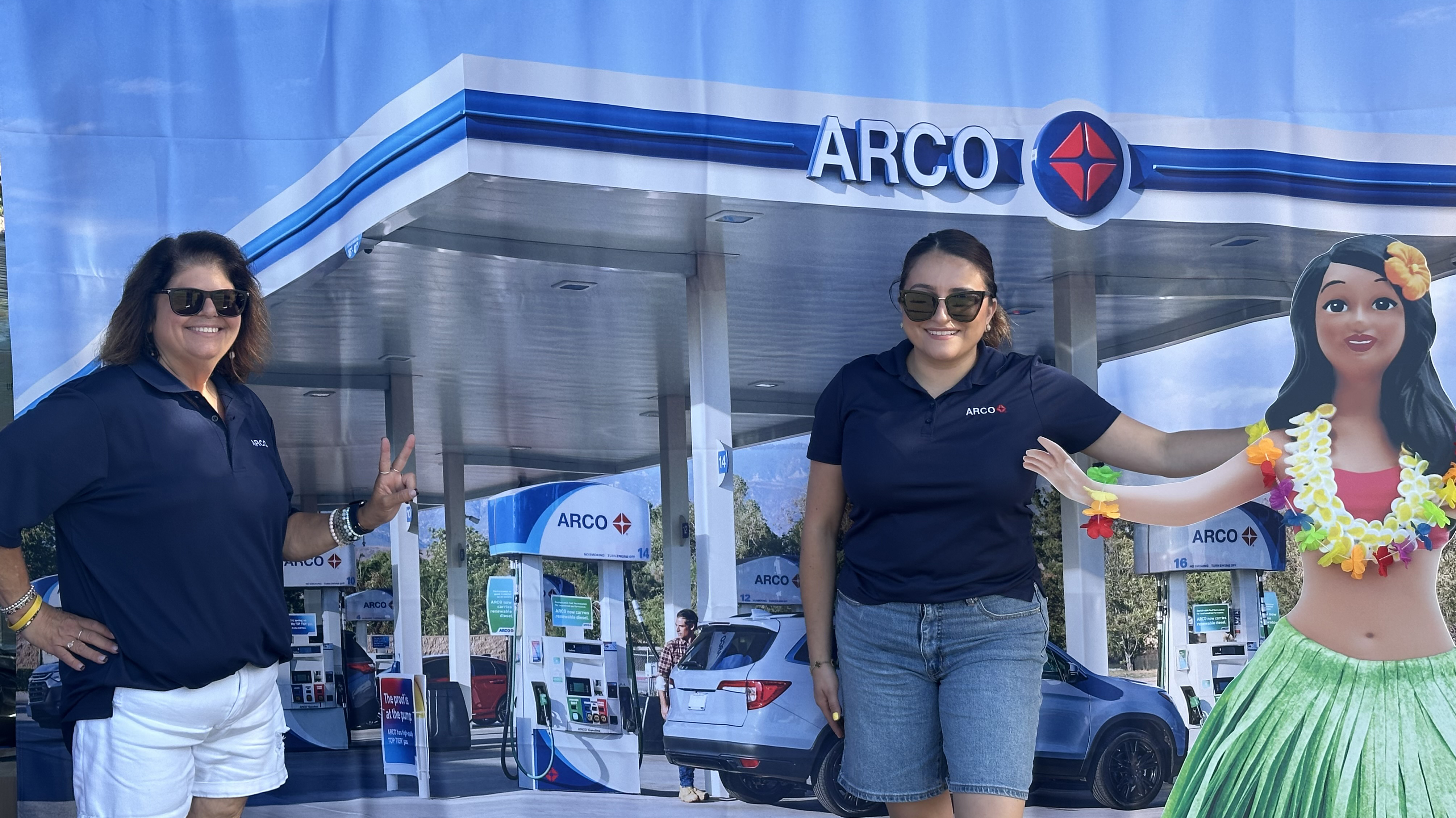 Two women stand in front of a backdrop with an ARCO gas station on it and the character Hanna in the foreground. 
