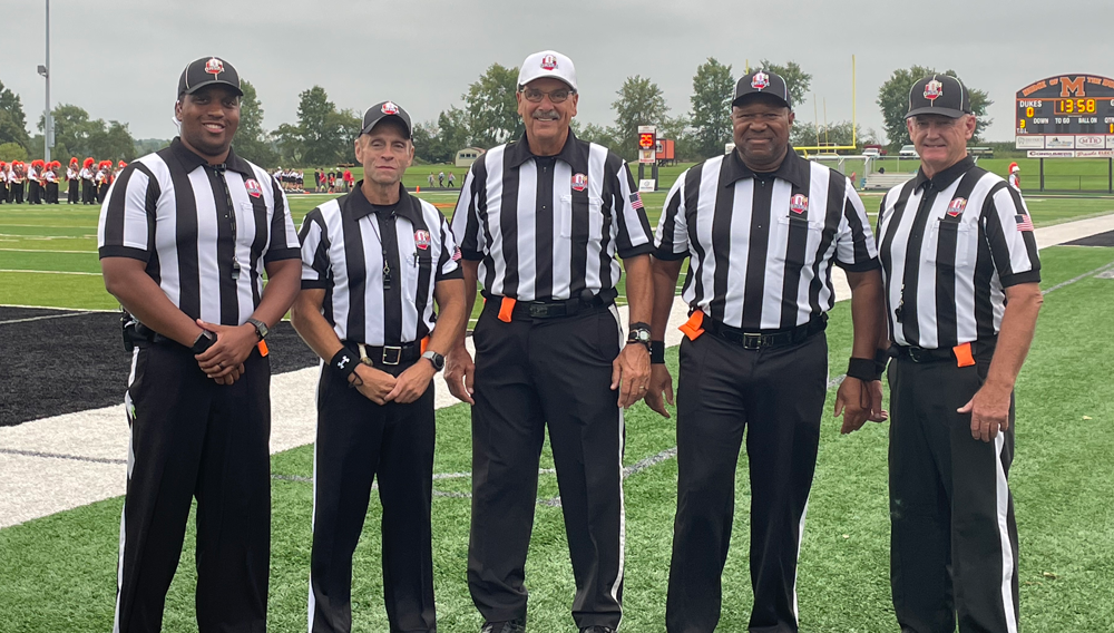 A group of five football referees in black and white striped shirts stand together on the field.
