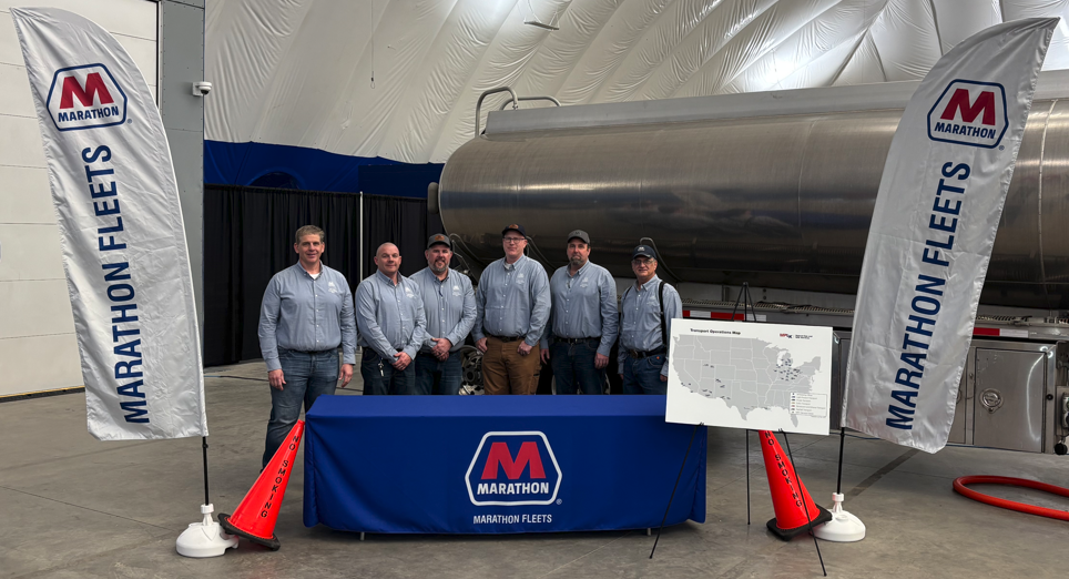  Employees in blue shirts stand in front of a tanker and between signs that read Marathon Fleets.