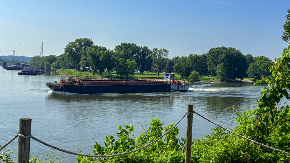 A boat on the water with blue skies and greenery in the foreground beside a roped fence.