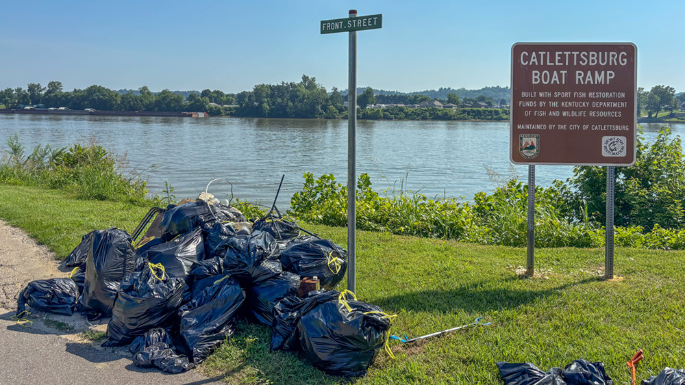 Black trash bags piled on the ground near a sign by the water.