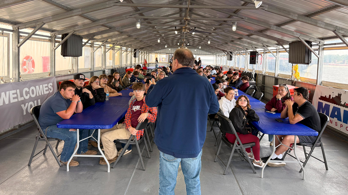 A man stands in front of rows of students sitting at tables on a boat.
