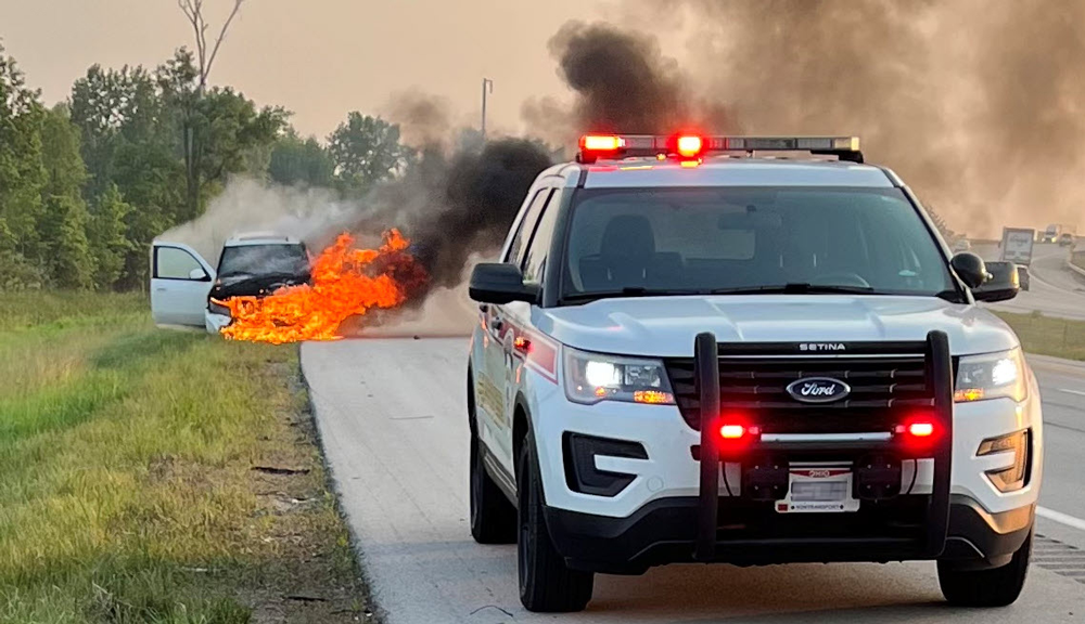 Police car with lights on in front of a white car on the side of the road with flames coming out of it. 