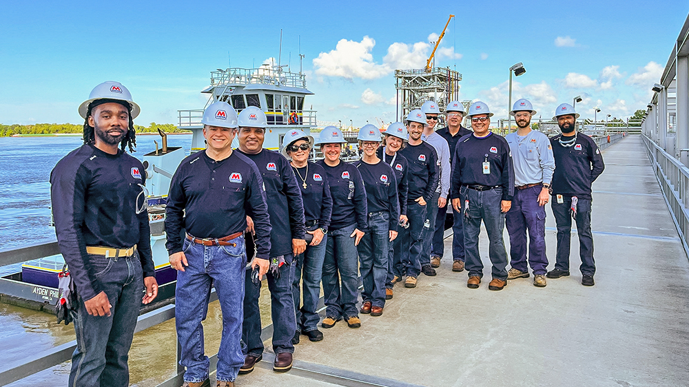  A group of people wearing hardhats stand on a dock in front of a boat in the water.