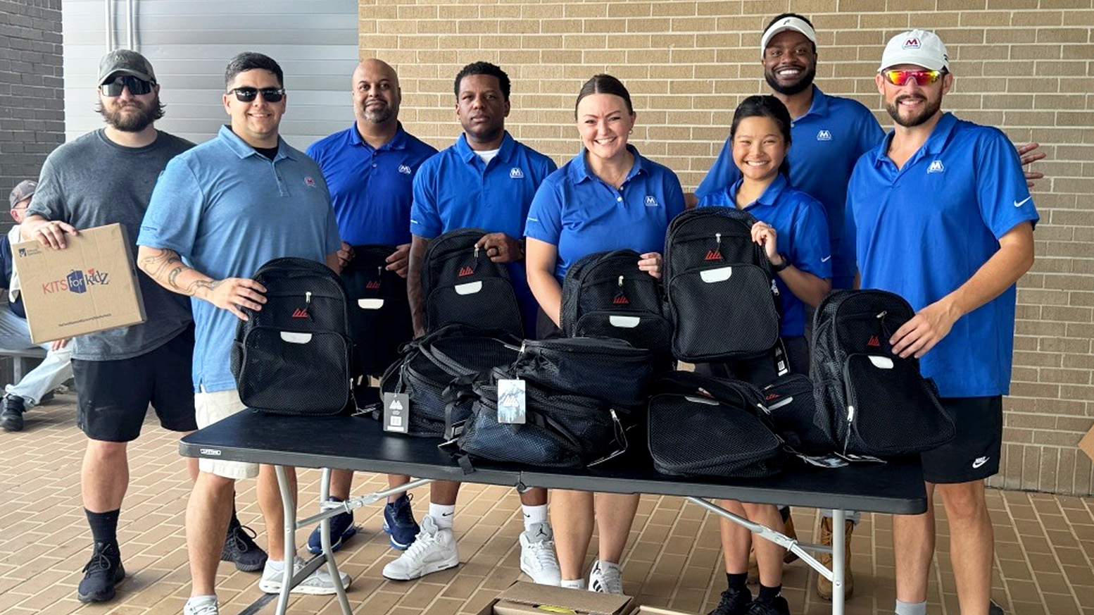 A group of people in blue shirts holding black backpacks on a table. A group of people in blue shirts holding black backpacks on a table.