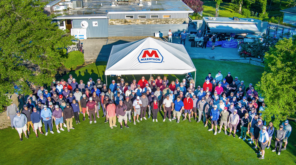 An aerial shot of a large group of people standing on a golf course near a white tent with a Marathon logo.