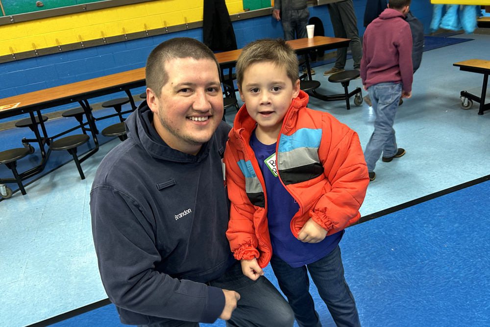An adult leans down and smiles at a child indoors after helping with a coat.
