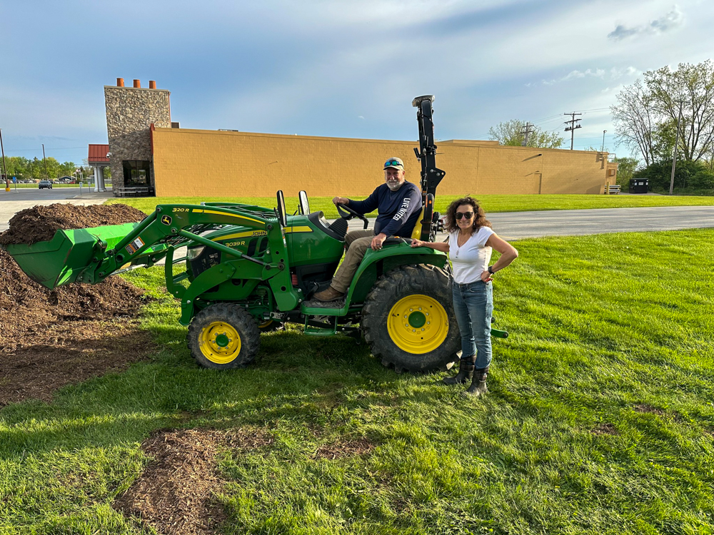 A woman stands beside a tractor while a man sits in the driver&rsquo;s seat, posing for a photo outdoors.