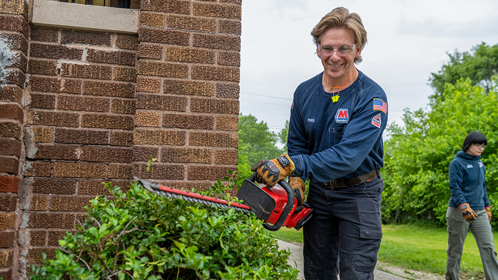 A volunteer holds a trimmer over a bush by a brick home.