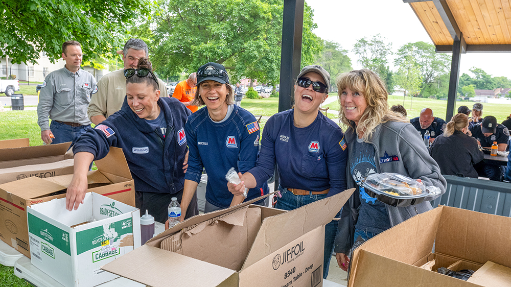 Group of volunteers smile under a pavilion with boxed lunches on a table.