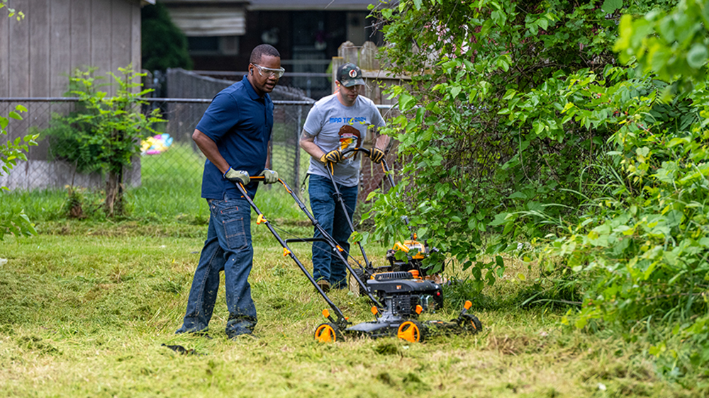 Two men each push a lawn mower through a yard.