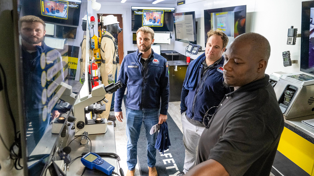 Three people look at a monitor on the wall inside a trailer filled with technology.