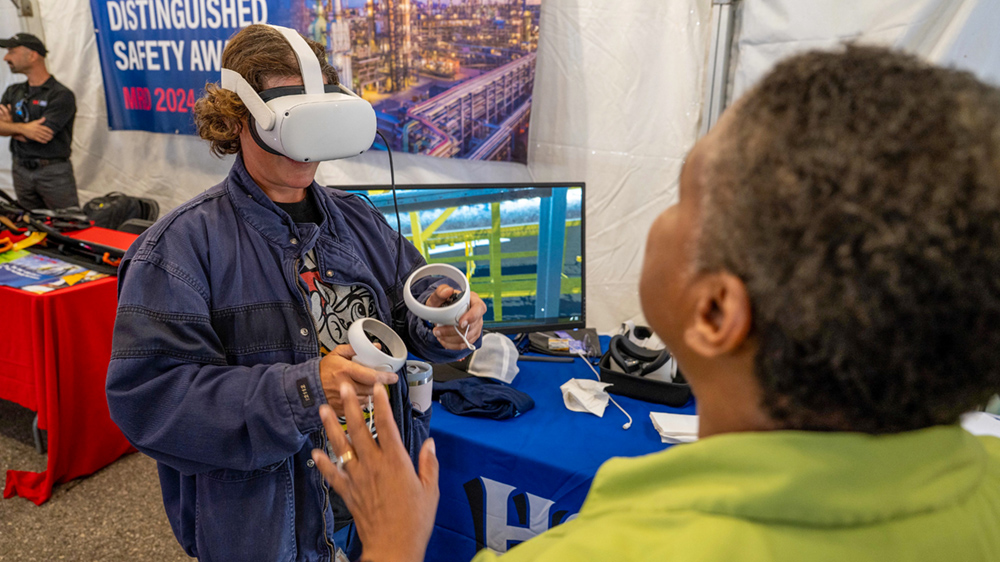 A person wearing virtual reality headset and holding remote controls in their hands by a display table.