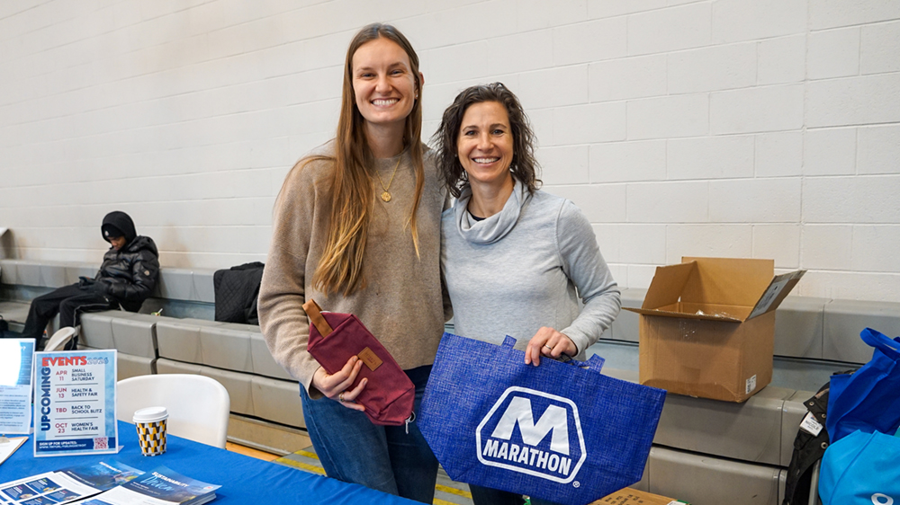 Two people hold up branded bags at a table with flyers and swag on it. 