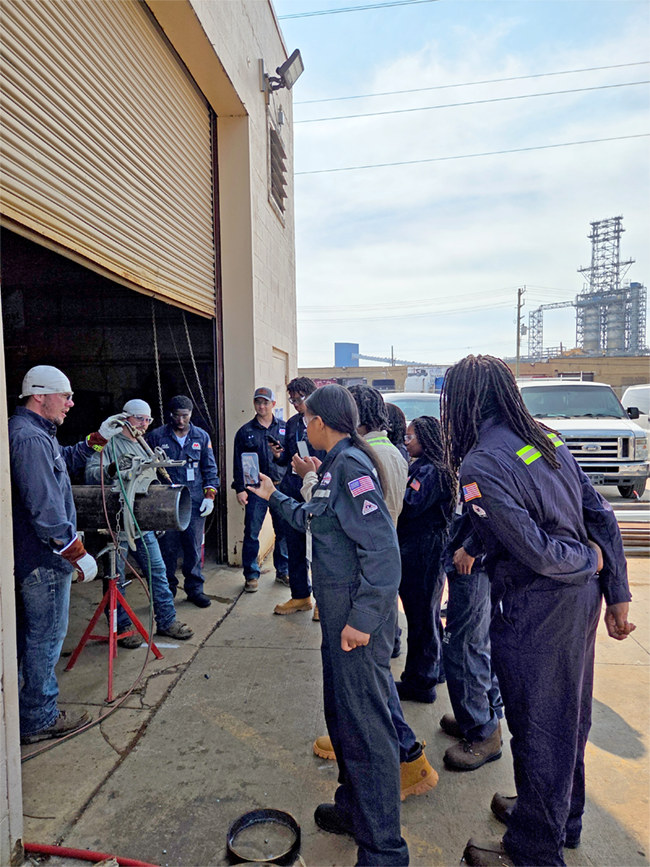 A group of students in blue protective coveralls stand outside a maintenance garage.