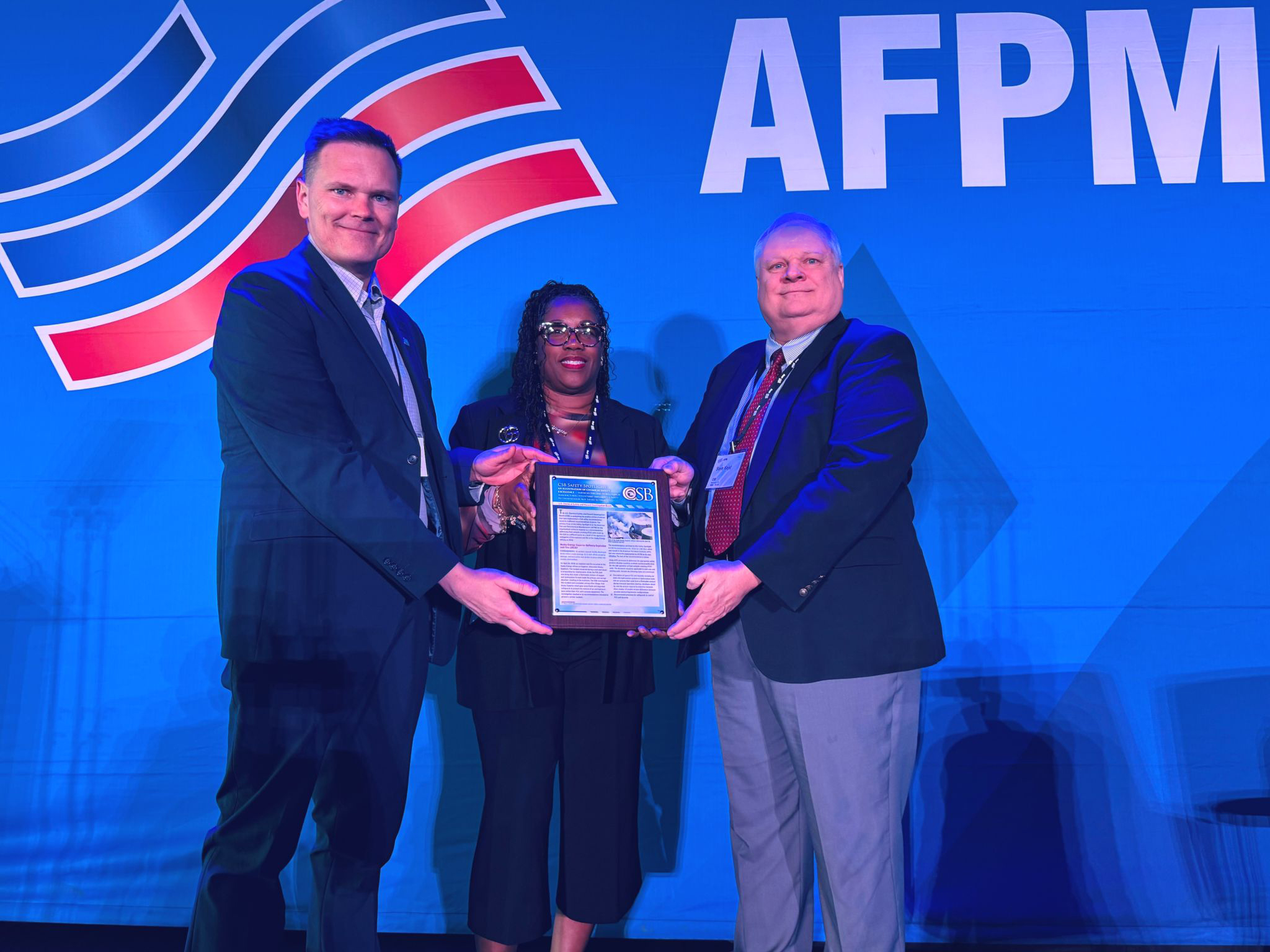 Three people hold a plaque on stage in front of a blue background that has the AFPM logo.