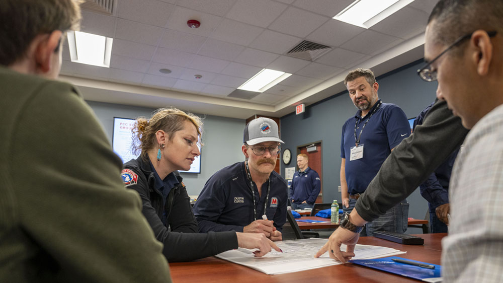 A group of people around a table discuss and point at a process control overview document. 