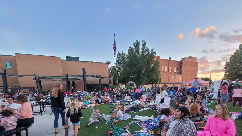 Hundreds of people sit in a grassy area of a public gathering space. 