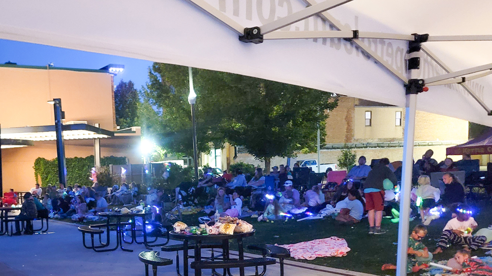 Hundreds of people sit outdoors at night in a grassy area of a public gathering space.