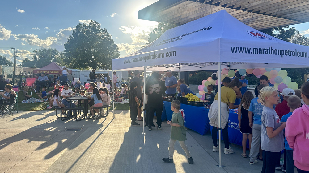 Dozens of people at tables and booths outside in a public gathering space. 