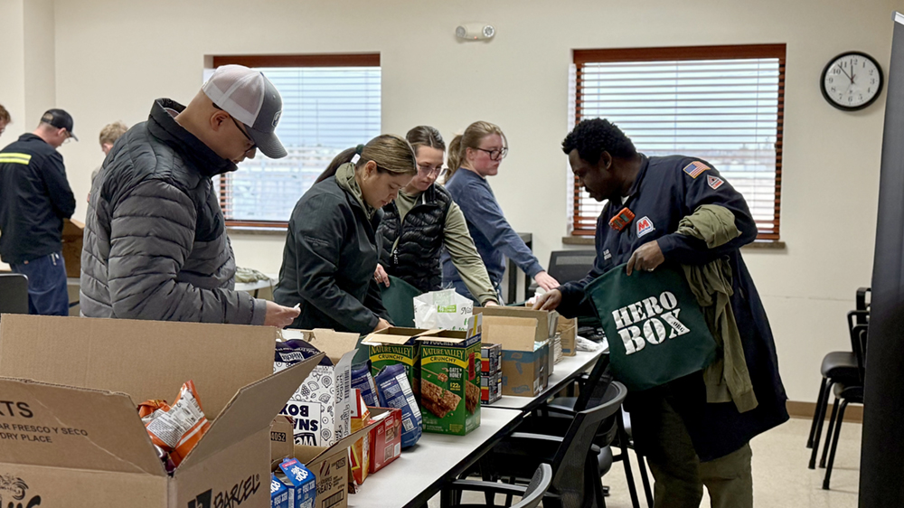 People work together around a table of supplies during a care package packing event in a large conference room.