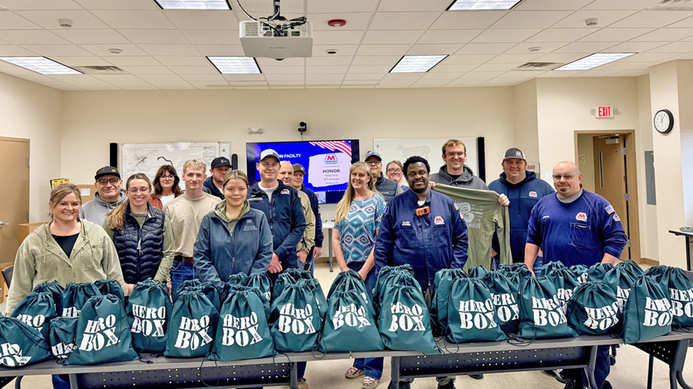 People stand behind a table, smiling, with green bags filled with donated items lined up in front of them.