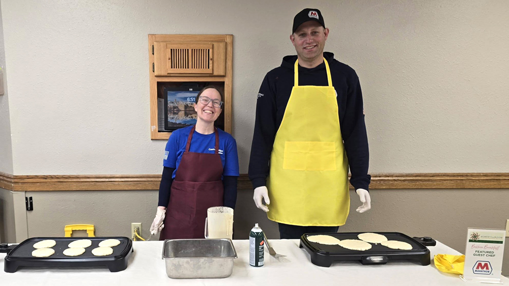 Two people stand behind a table where they are making breakfast at a community event.