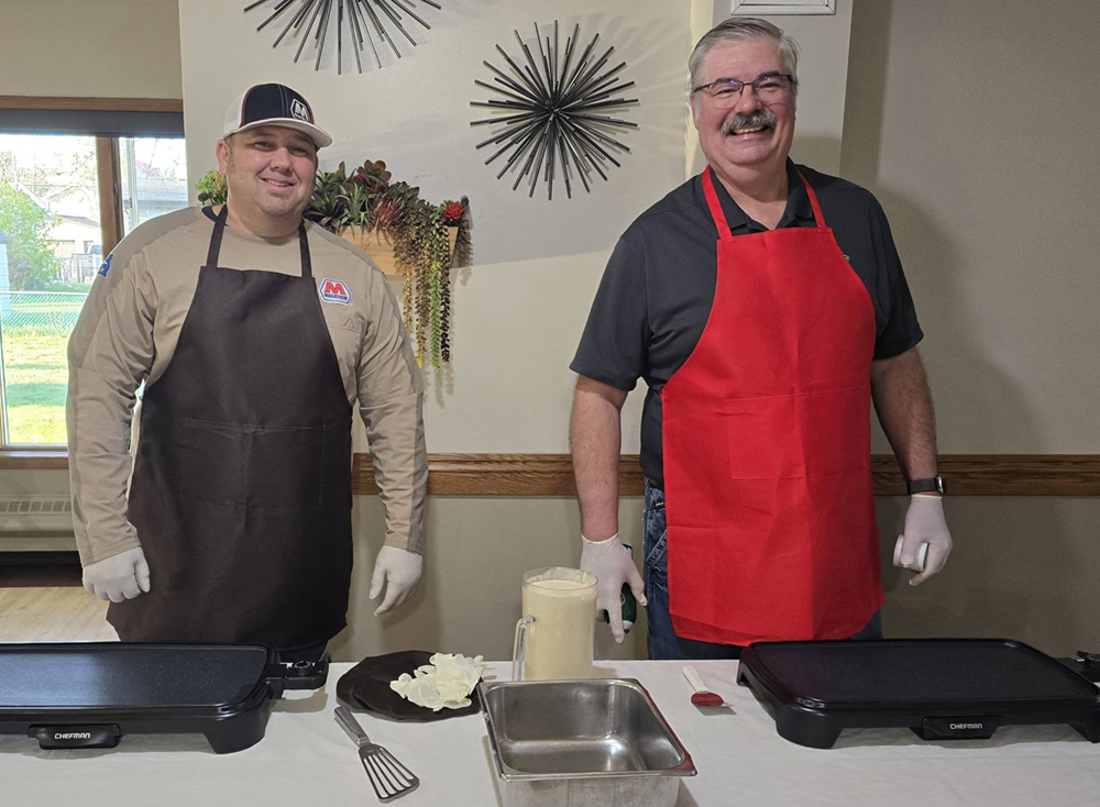 Two men wearing aprons pose for a photo at a community event.