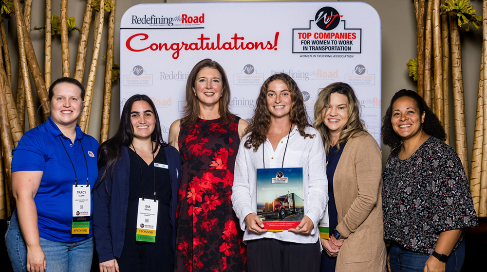 5 women are standing in front of a sign and holding awards.