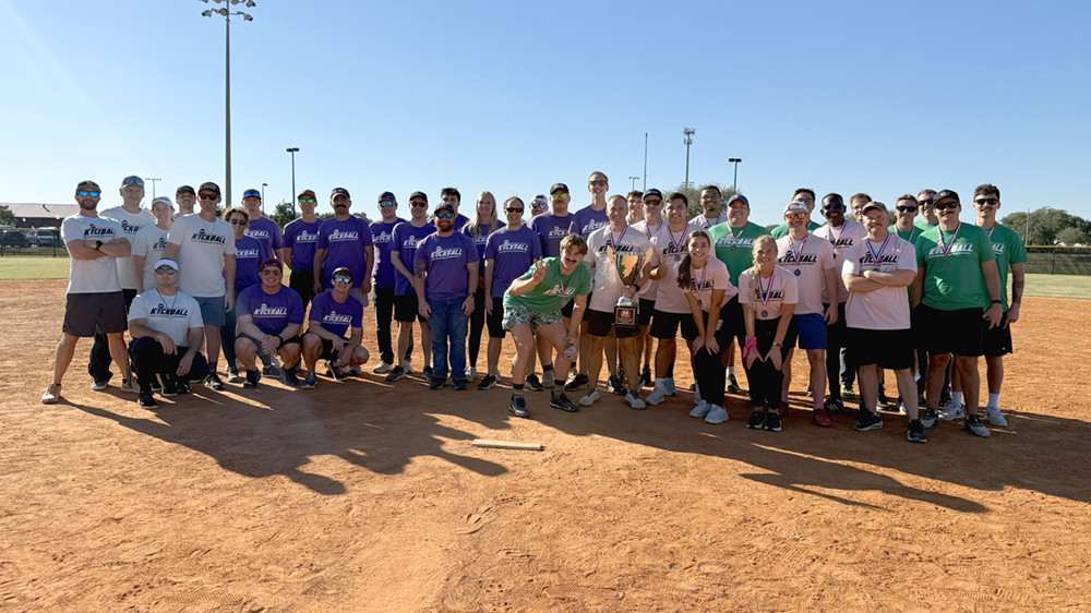 A large group of people standing together on a baseball-style field, with blue skies and tall light poles in the background.