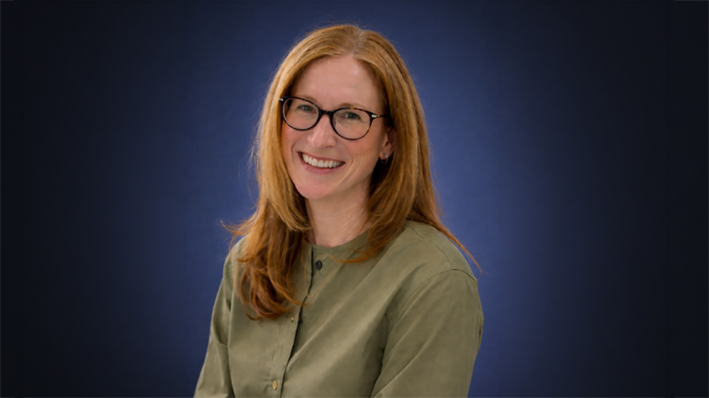 A photo of Heather Vail looking at the camera and standing in front of a grey background.