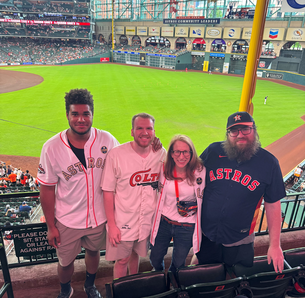 Four people stand together in the stands at a baseball game.