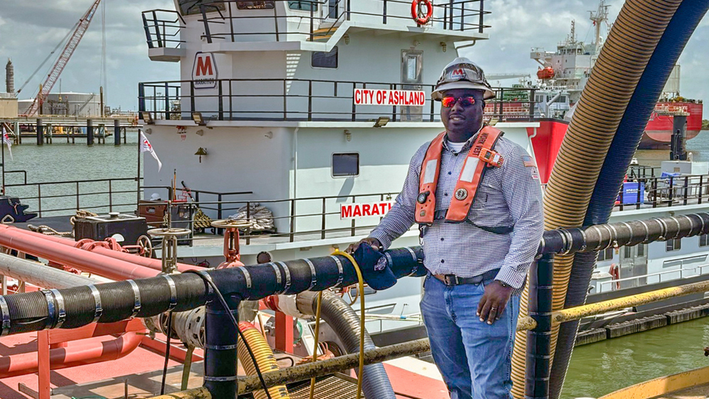 Man wearing safety gear stands on the shore in front of a vessel in the water.