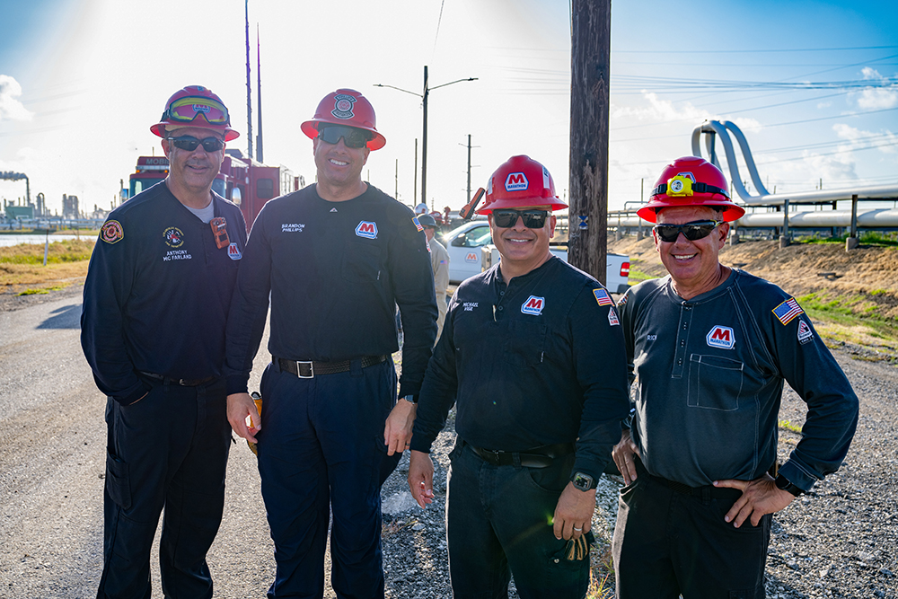 Four people wearing hard hats stand shoulder to shoulder outside an industrial complex.