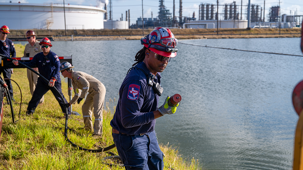 People in safety gear move a large water hose along a waterway during an emergency response drill at a Texas refinery.