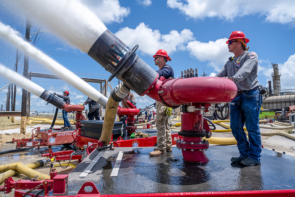 People wearing hard hats stand behind large water hoses aimed at a crude oil storage tank the size of a football field.