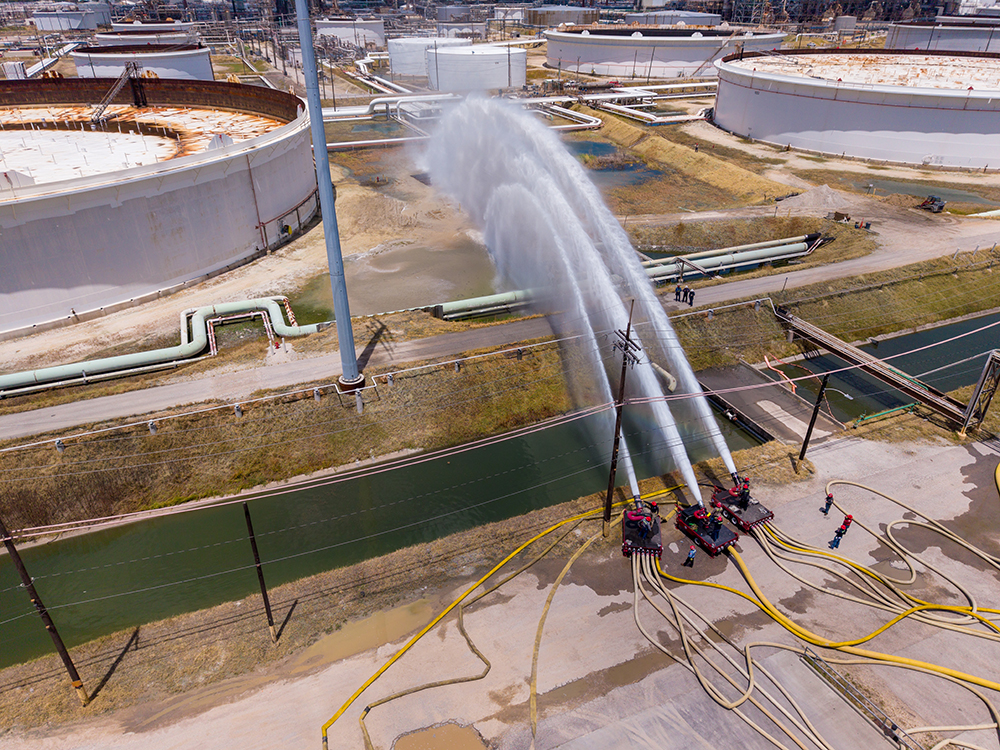 Emergency crews spray water from large hoses toward a crude oil storage tank during a full-scale emergency response drill at a refinery.