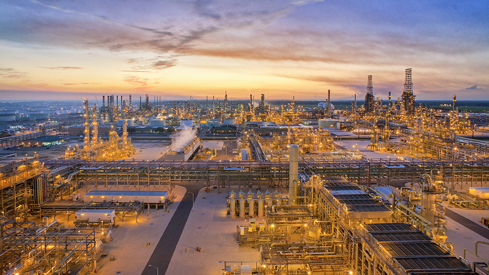 Aerial view of an illuminated refinery at sunset beneath a colorful, partly cloudy sky.