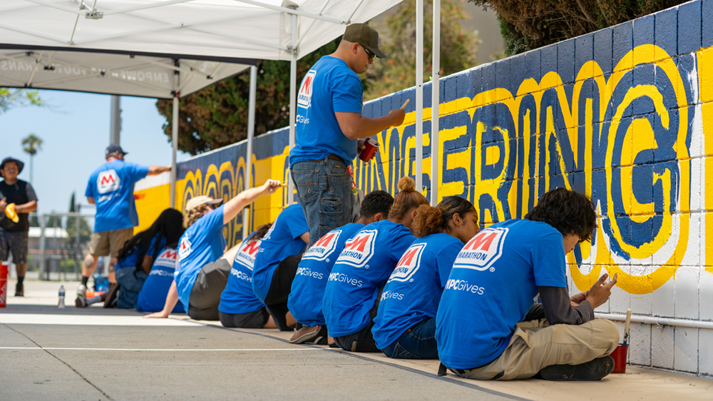 : A line of people in blue volunteer shirts paint a mural wall.