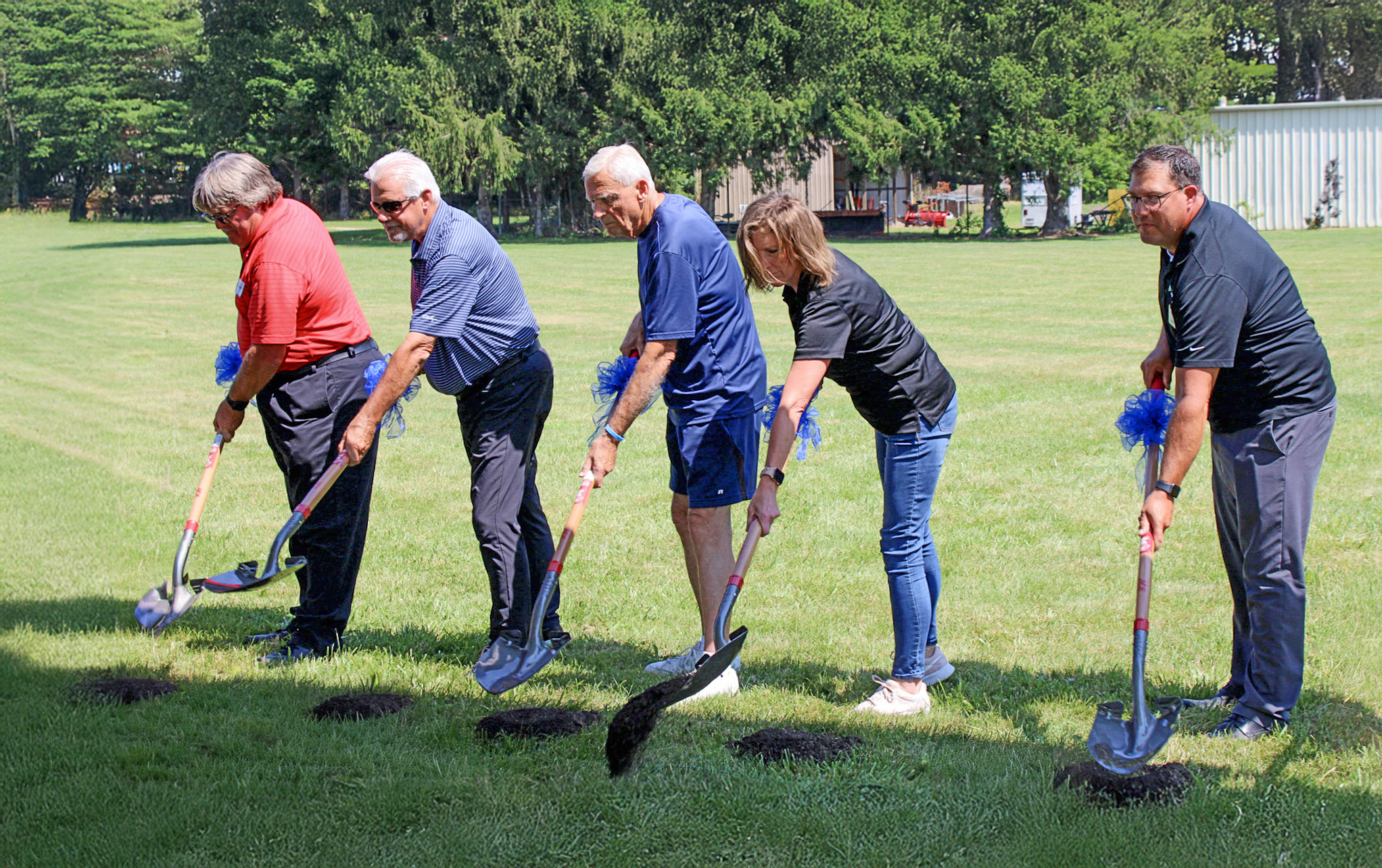  Five people lined up holding shovels and digging in the ground.