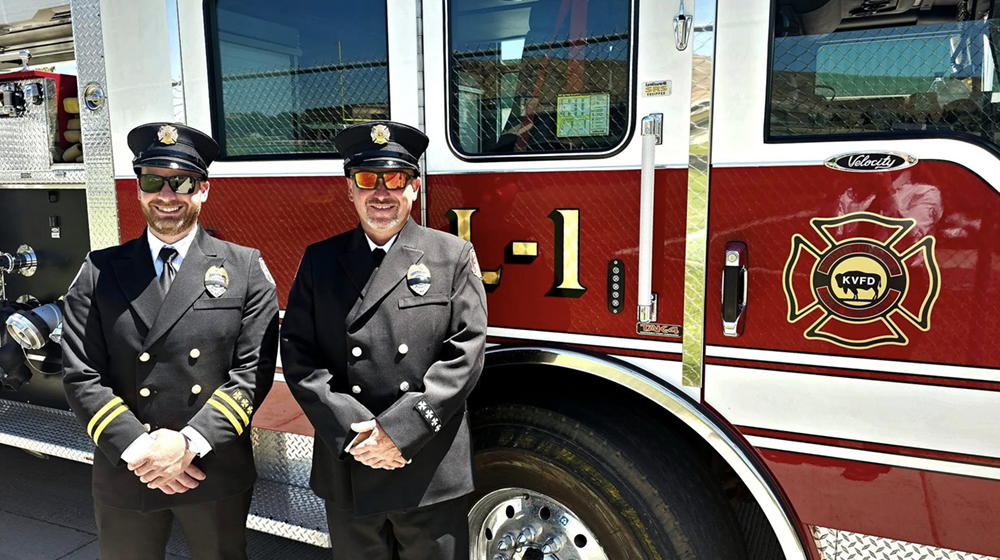Two firefighters in uniform stand in front of a fire truck.